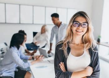 Blonde female executive posing with smile and arms crossed during brainstorm with managers. Indoor portrait of european student spending time in hall with asian and african friends.